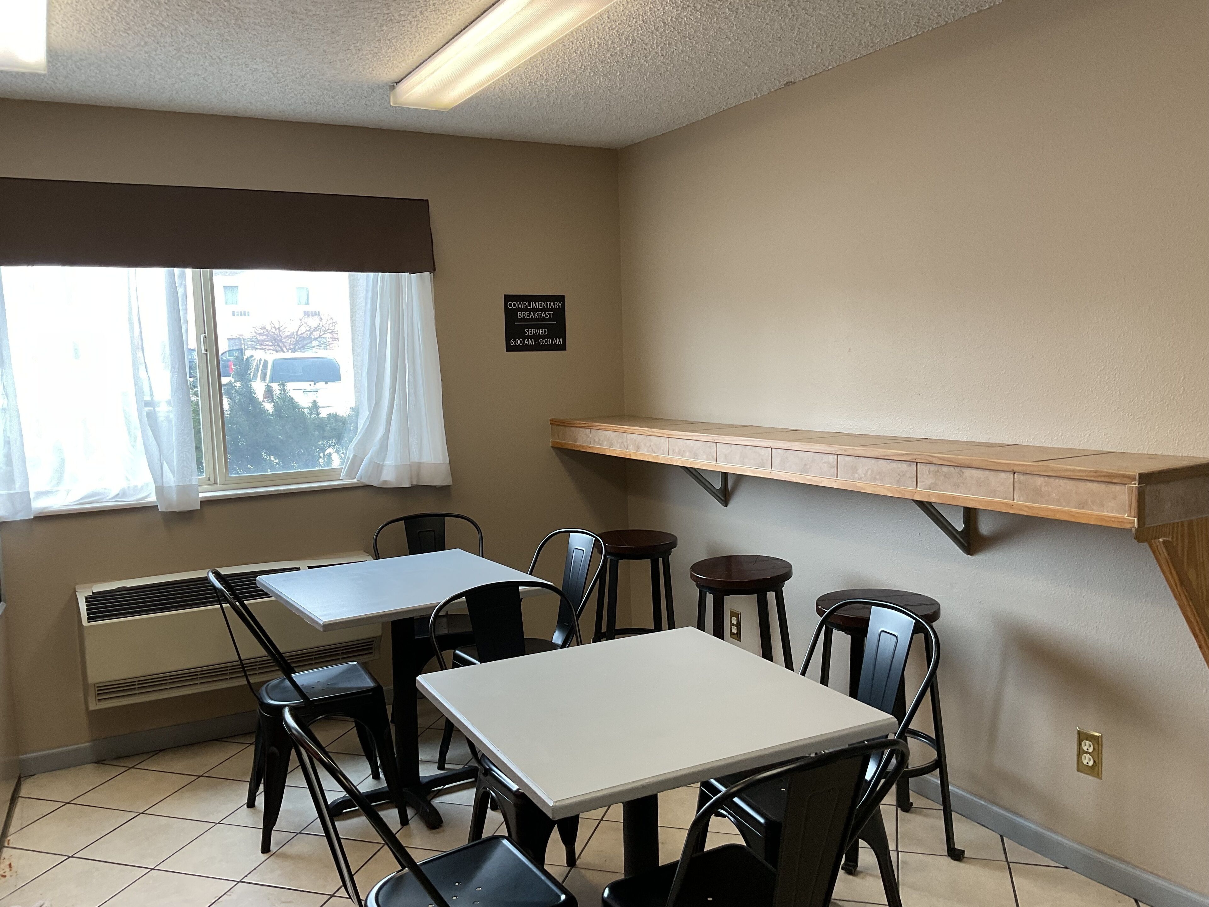 A small dining area with a table and chairs, a window with curtains, and a sign indicating complimentary breakfast hours.