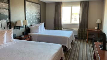 A hotel room with two beds, a desk, a chair, a lamp, a window with curtains, and a wall-mounted air conditioner.
