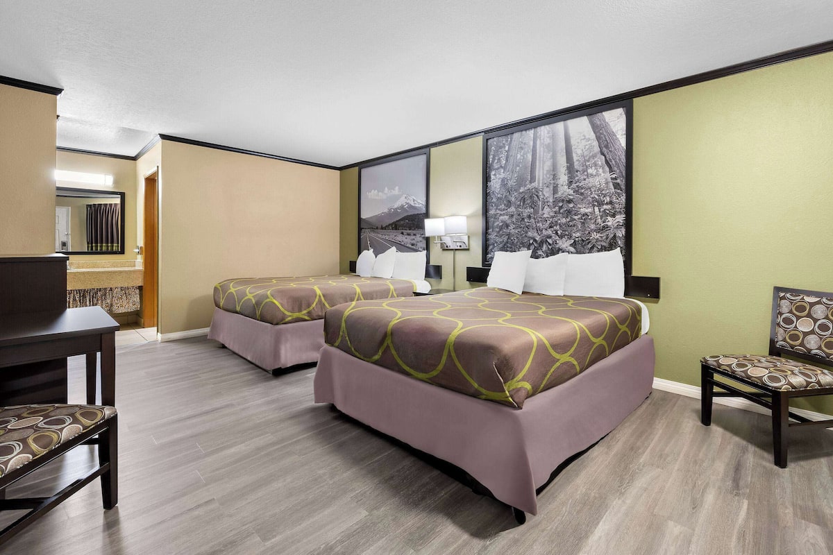 A hotel room with two beds, a desk, a chair, and a large window with a view of a forest.