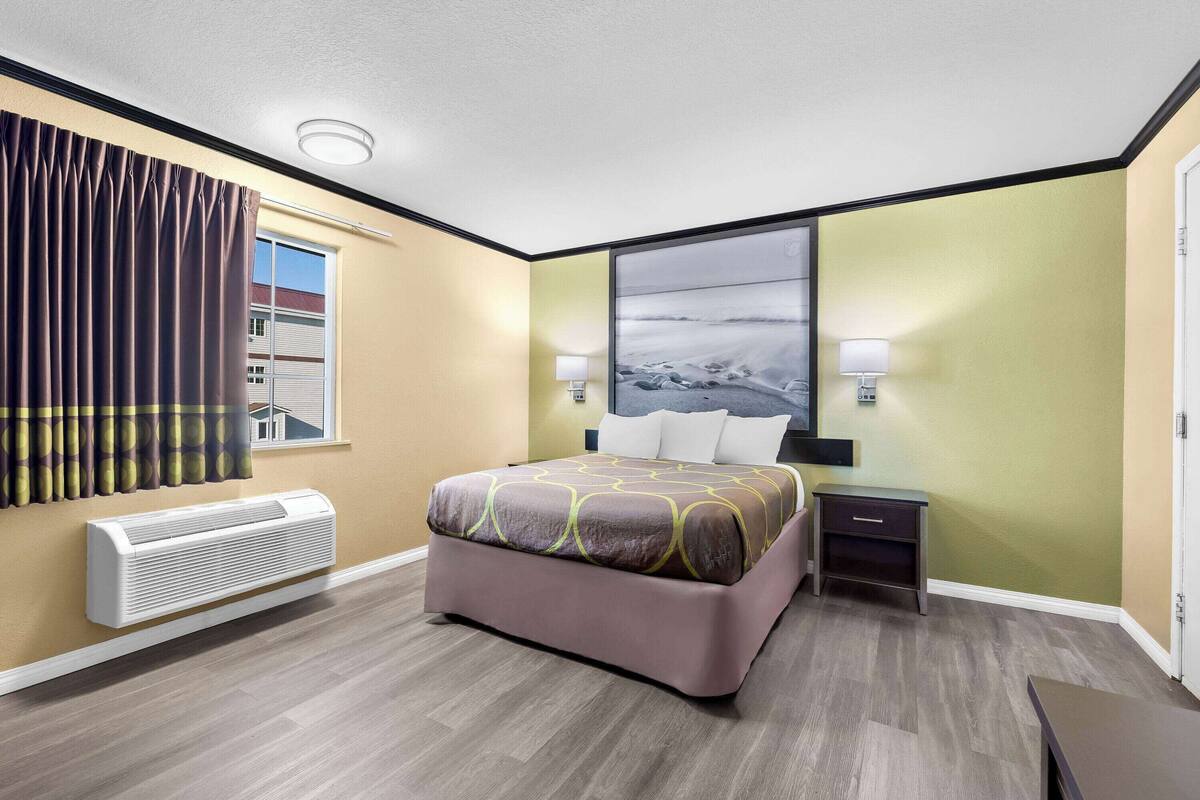 A hotel room with a bed, bedside tables, a window with curtains, a wall-mounted heater, and a large painting on the wall.