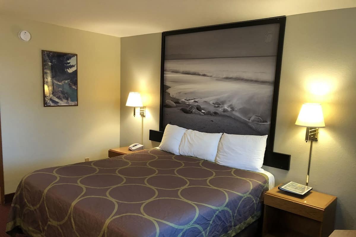 A hotel room with a bed, two bedside lamps, a nightstand, a phone, a framed picture on the wall, and a large window with a view of the beach.