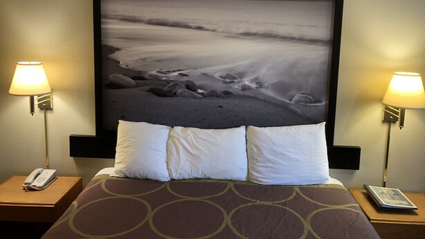 Ein Hotelzimmer mit einem Bett, zwei Nachttischlampen, einem Telefon und einem gerahmten Foto einer Strandansicht.