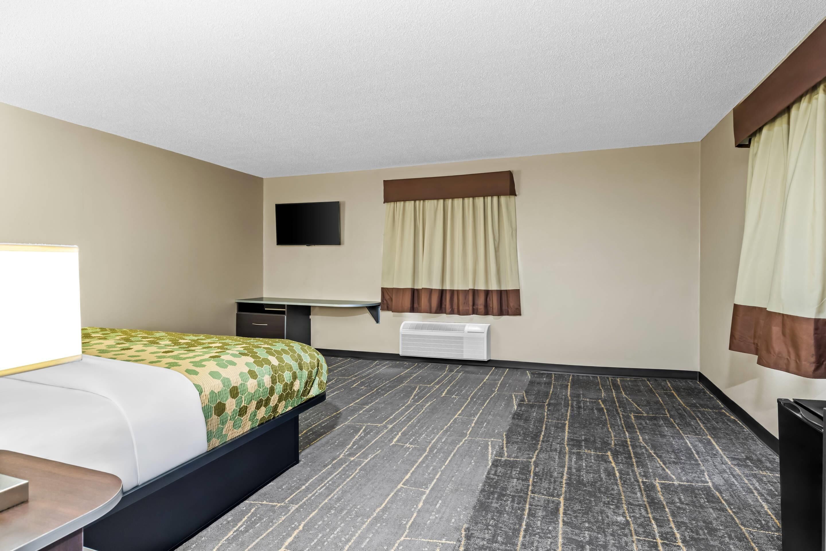 A hotel room with a bed, a desk, a flat-screen TV, a window with curtains, and an air conditioning unit.