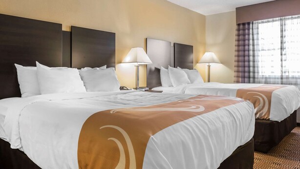 Habitación de hotel con dos camas, cada una con ropa blanca y detalles marrones, y cabeceros oscuros. Hay dos lámparas de noche y una ventana con cortinas.