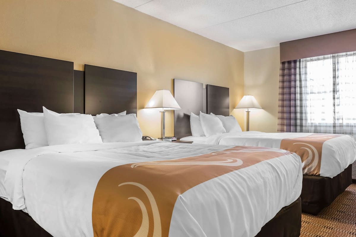 A hotel room with two beds, each with white bedding and brown accents, flanked by dark headboards. There are two bedside lamps and a window with curtains.