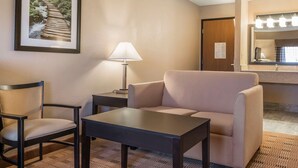 Habitación de hotel con sofá, una silla, una mesita, una lámpara, un cuadro en la pared y un baño con espejo e iluminación.