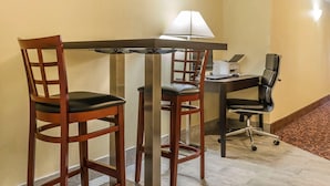 Habitación de hotel con una mesa de madera, dos sillas de madera, una silla de oficina negra, un escritorio con lámpara y una alfombra estampada.