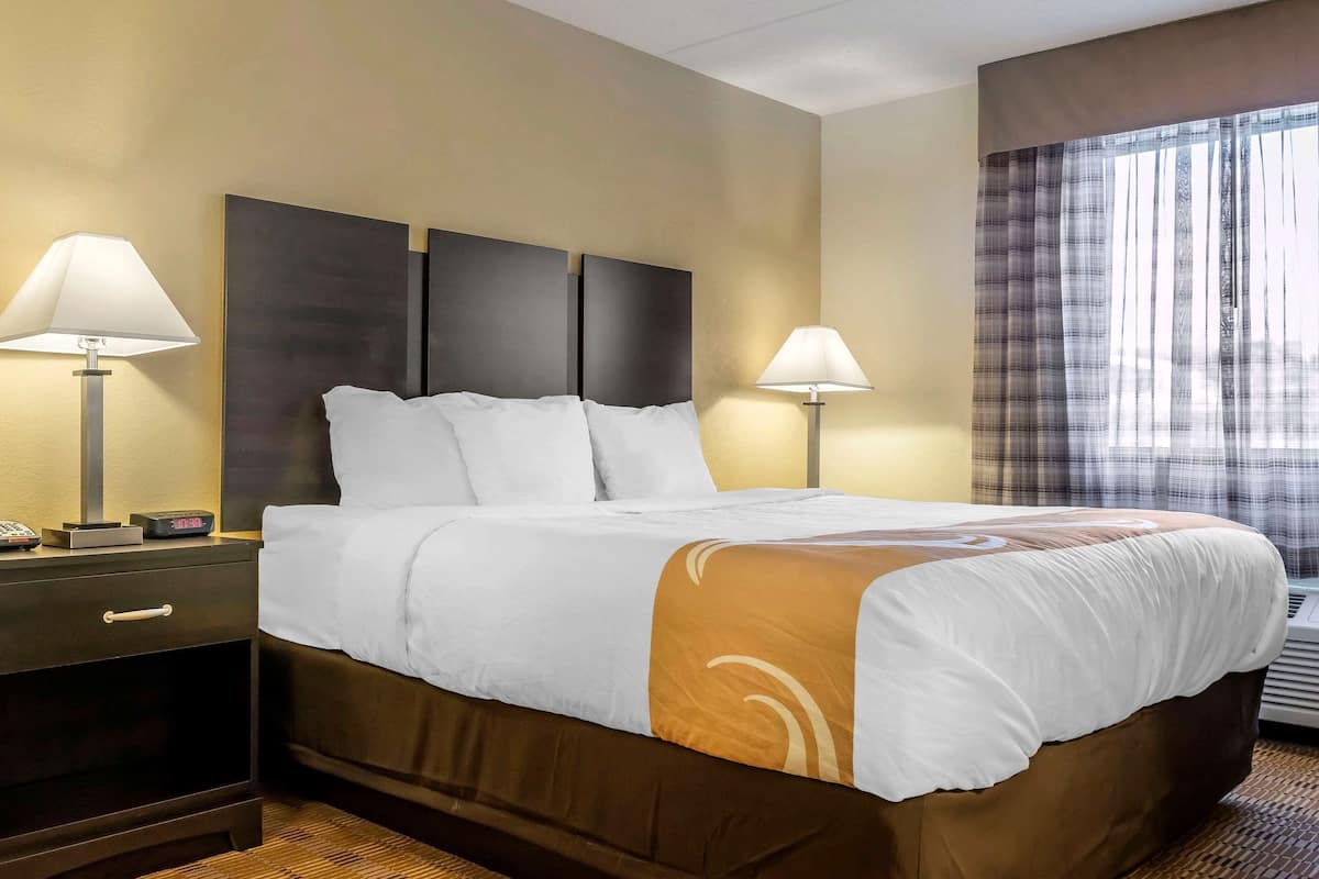 A hotel room with a large bed, two bedside tables with lamps, a headboard, and a window with curtains.