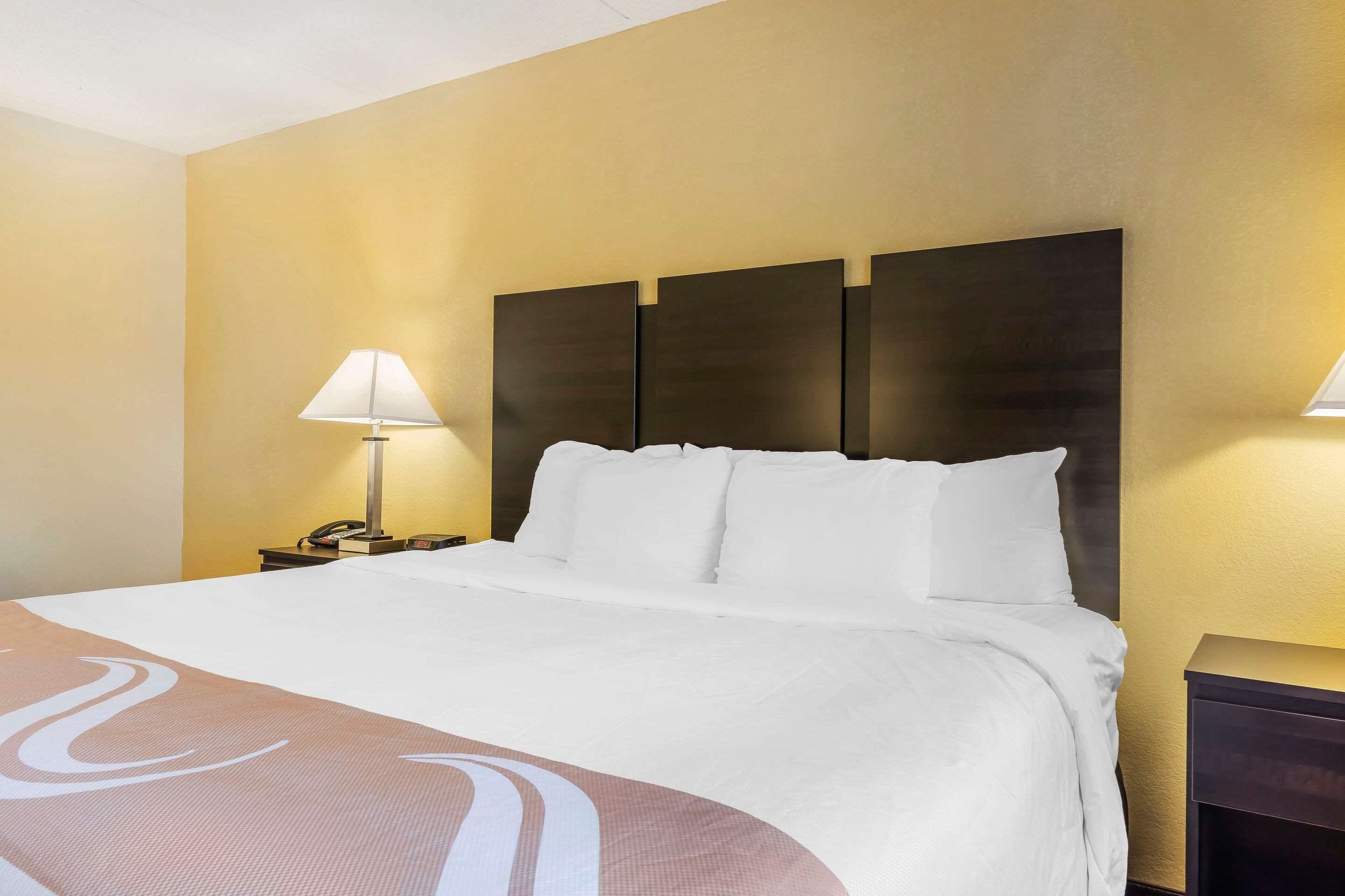 Habitación de hotel con una cama grande, una mesita de noche con lámpara y un cabecero fijado a la pared.