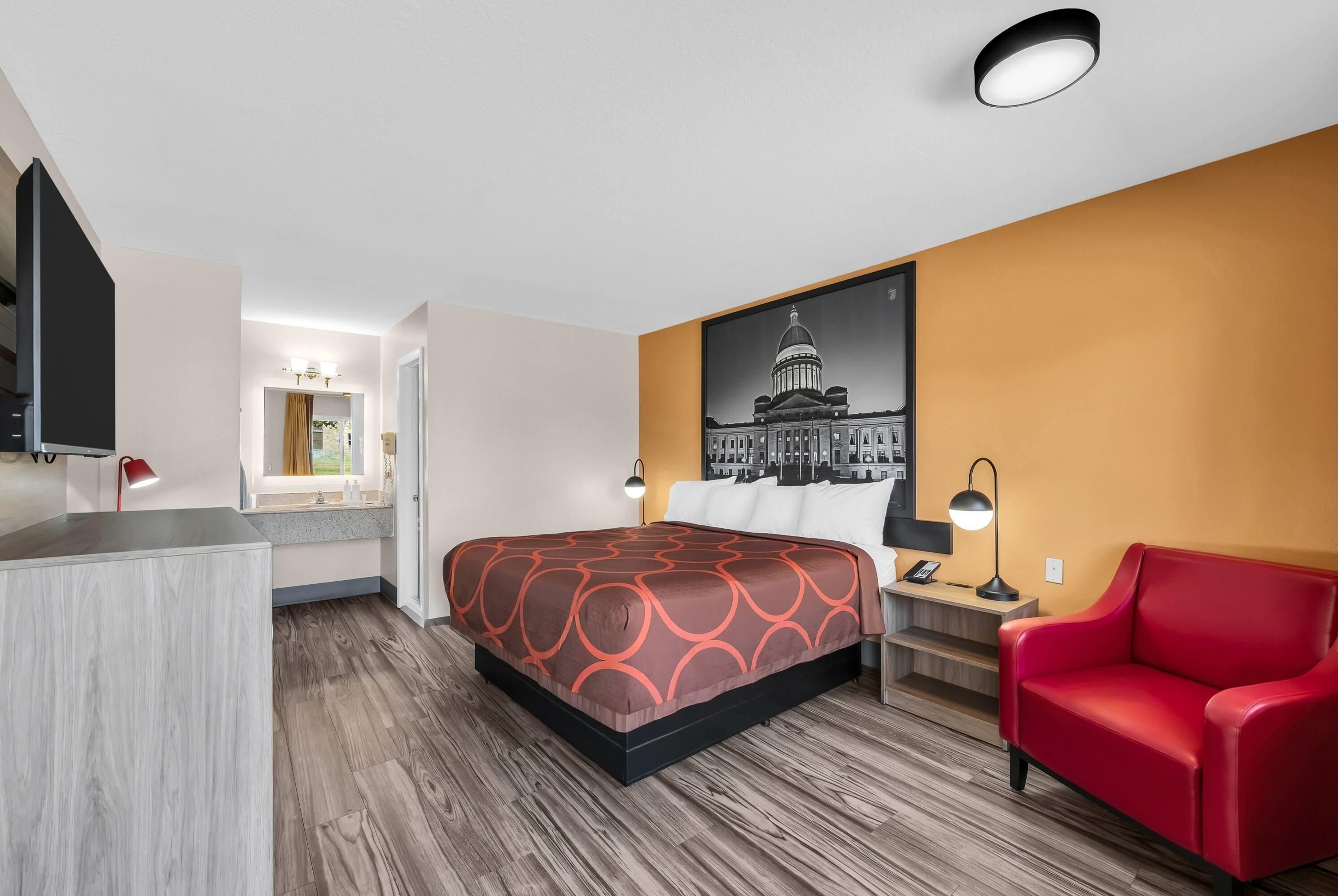 A hotel room with a bed, a red chair, a desk with a lamp, a bathroom, and a wall-mounted TV.