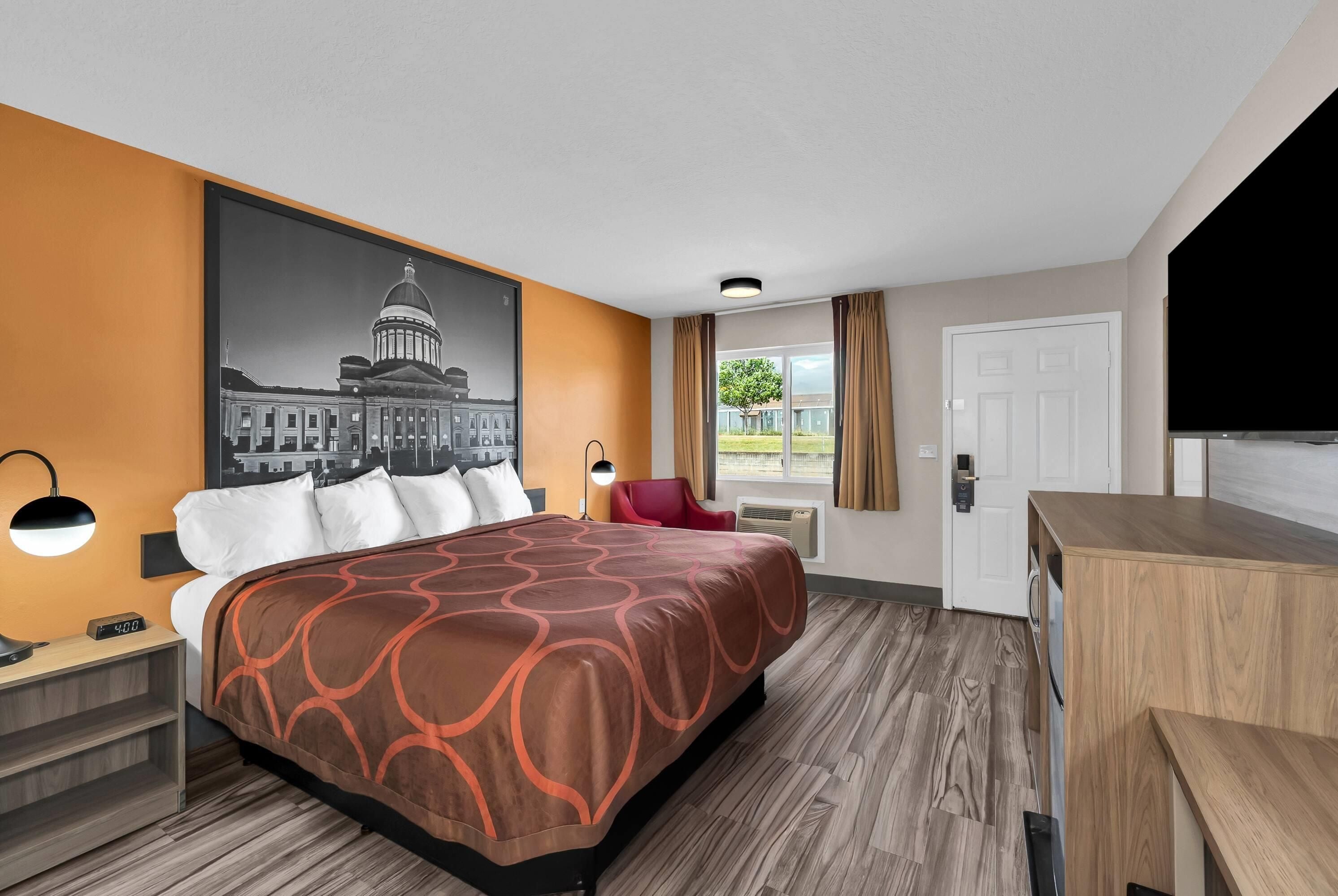 Ein Hotelzimmer mit einem großen Bett, einem Schreibtisch, einem Sessel, einem Fernseher, einem Fenster mit Vorhängen und einer Wand-Klimaanlage.