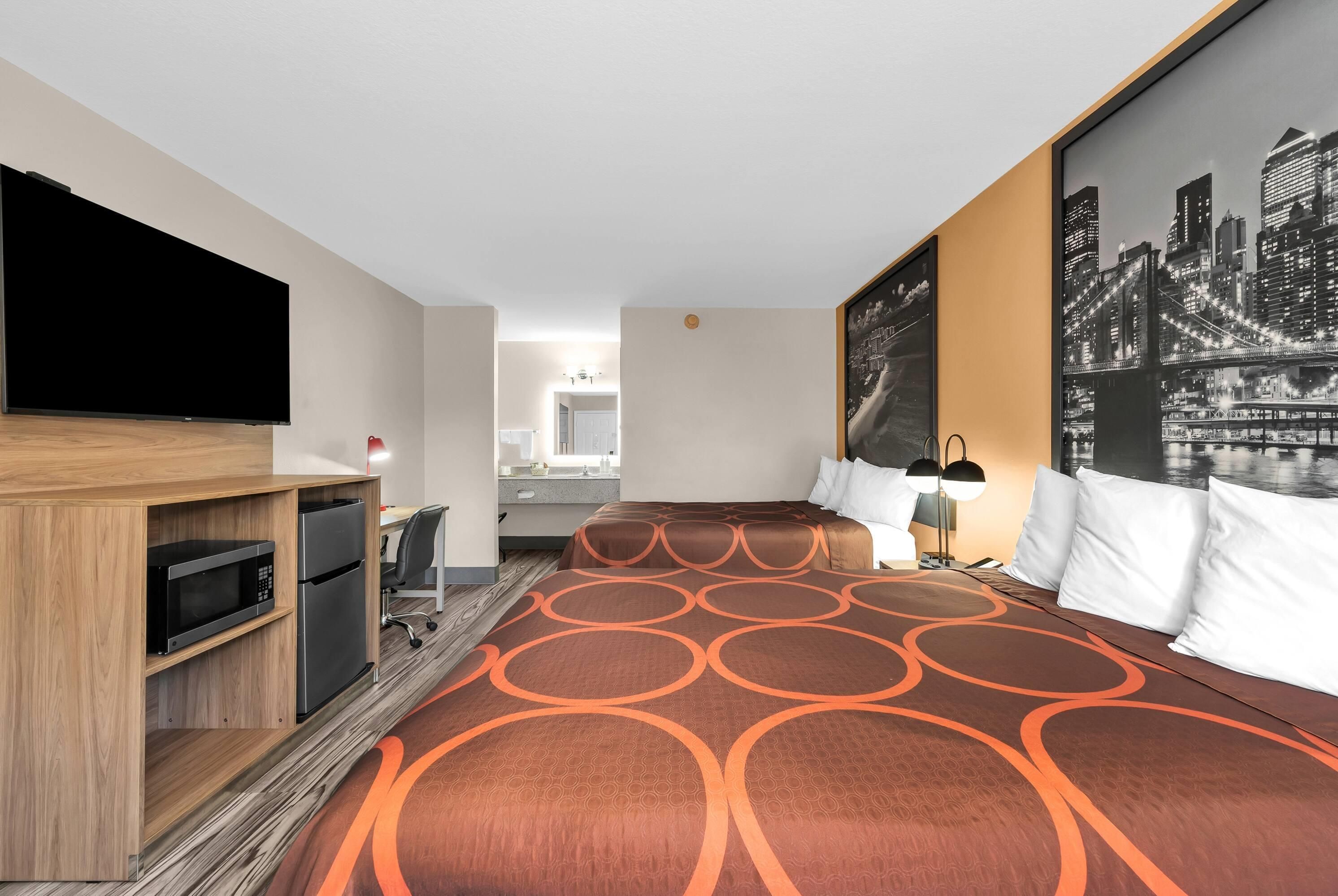 Une chambre d’hôtel équipée d’un lit, d’une télévision, d’un bureau, d’un réfrigérateur et d’une salle de bain, visible à travers une porte ouverte.