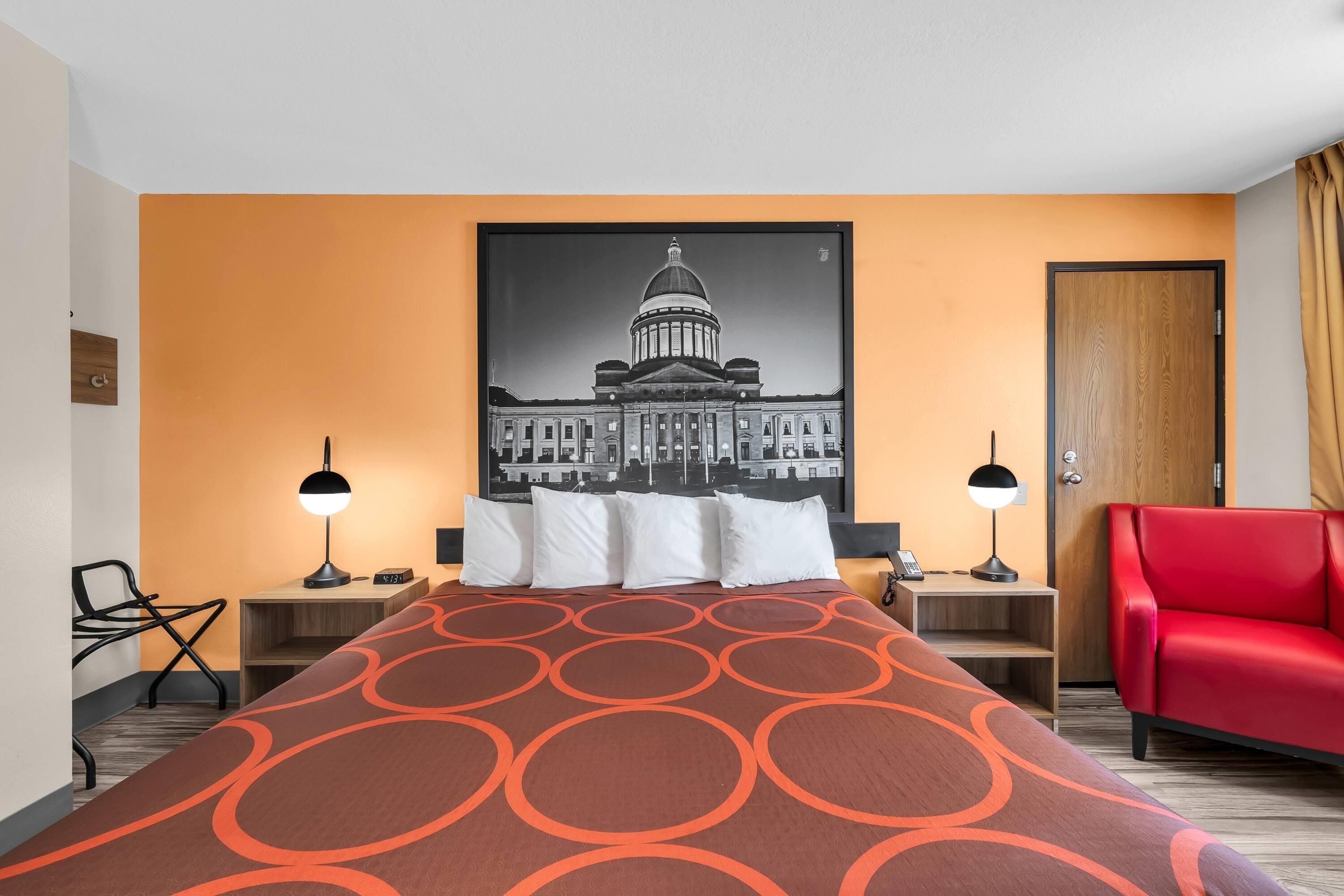 Ein Hotelzimmer mit einem Bett, zwei Nachttischen, einem roten Sessel, einer Wandlampe und einem Bilderrahmen mit einem Gebäude.