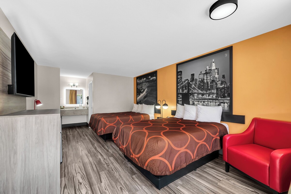 Habitación de hotel con dos camas, una silla roja, un cuadro de paisaje urbano y un televisor.