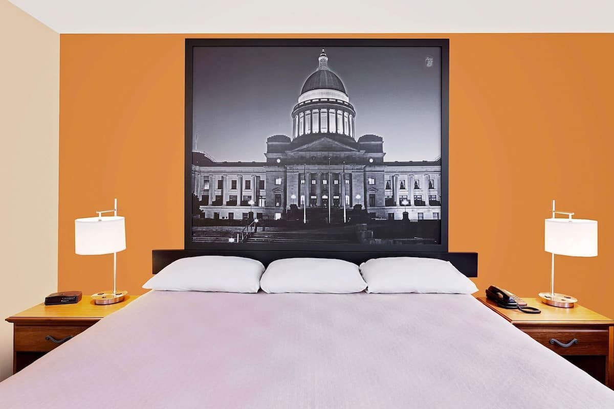 A hotel room with a large bed, two bedside tables, a nightstand, a framed picture of a building, and two lamps.