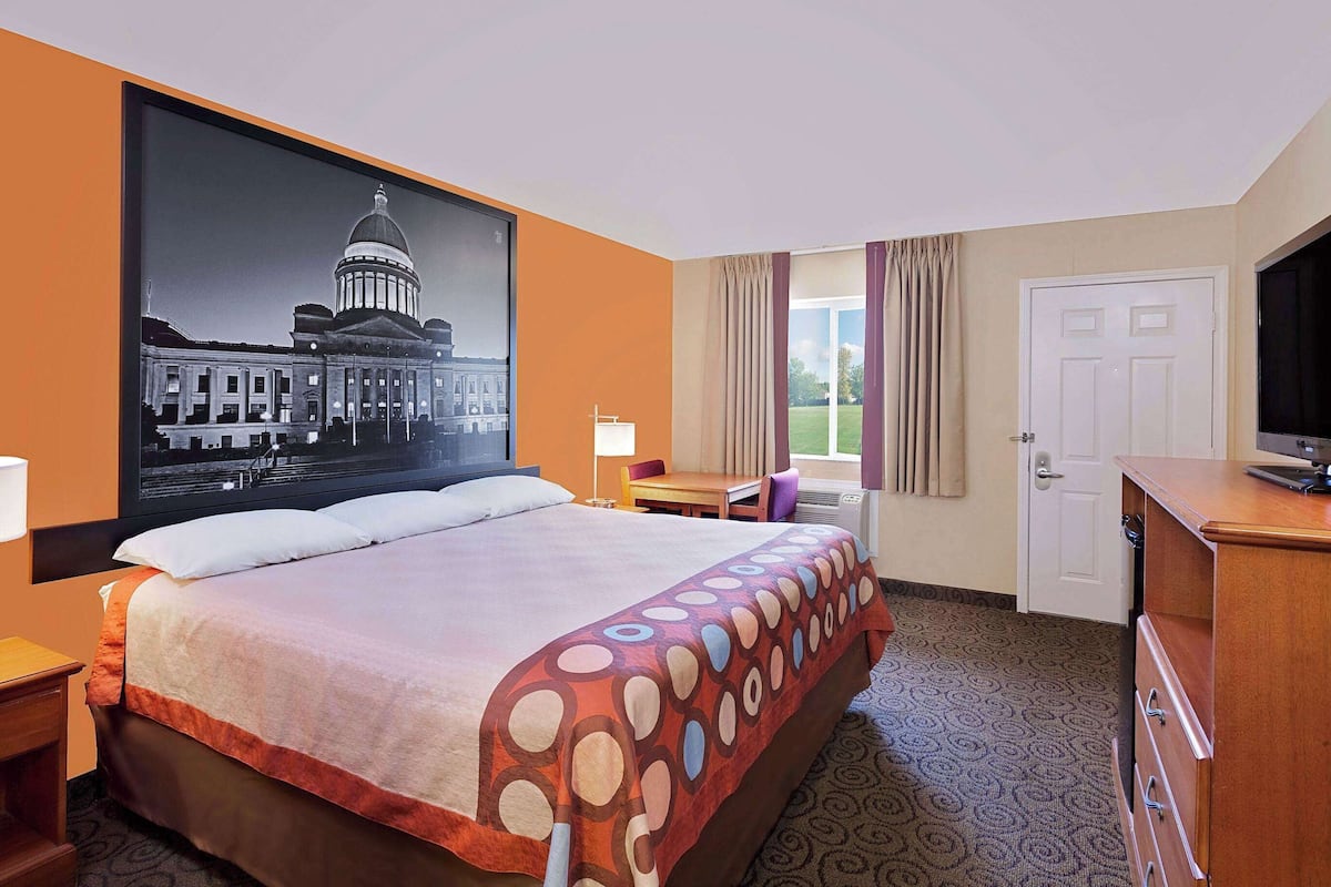 A hotel room with a large bed, a desk, a television, and a painting of a building.