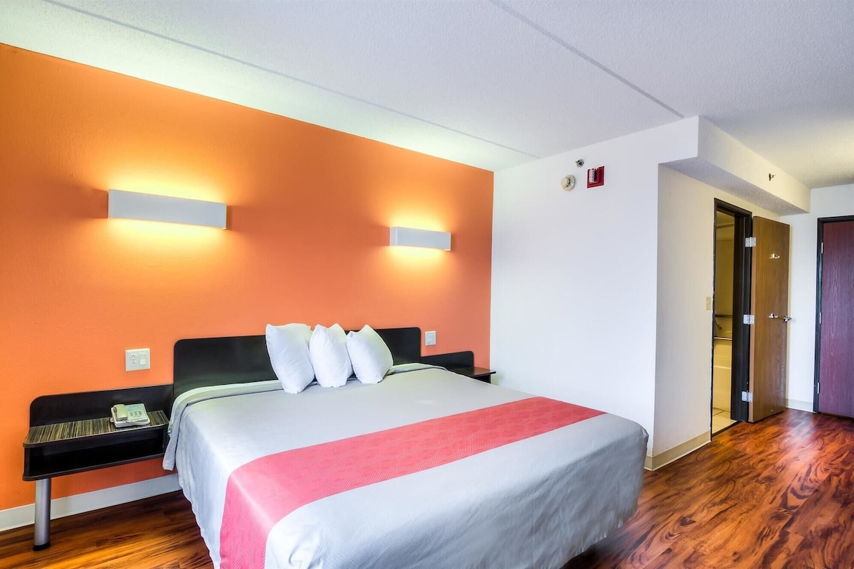 A hotel room with a bed, a bedside table, a nightstand, and a wall-mounted light fixture.