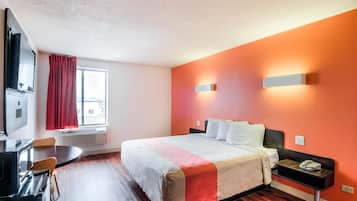 A hotel room with a bed, a desk, and a chair. There is a window with curtains, a wall-mounted TV, and a wall-mounted light fixture.