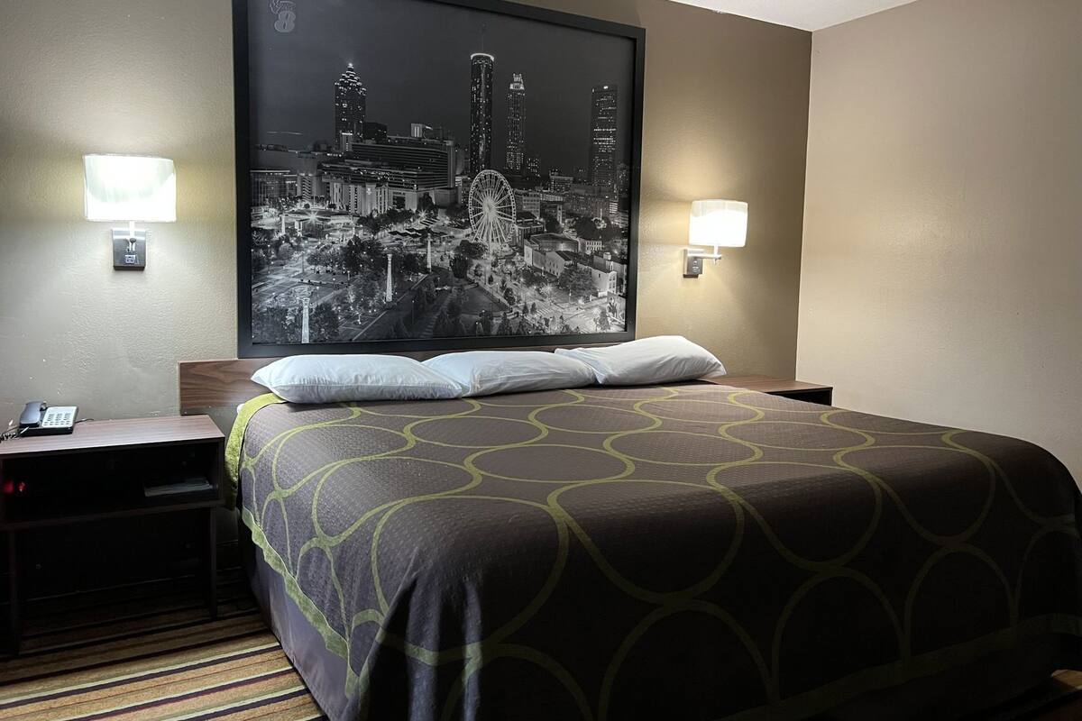 A hotel room with a large cityscape artwork on the wall, a bed with a patterned bedspread, bedside lamps, and a nightstand with a phone.