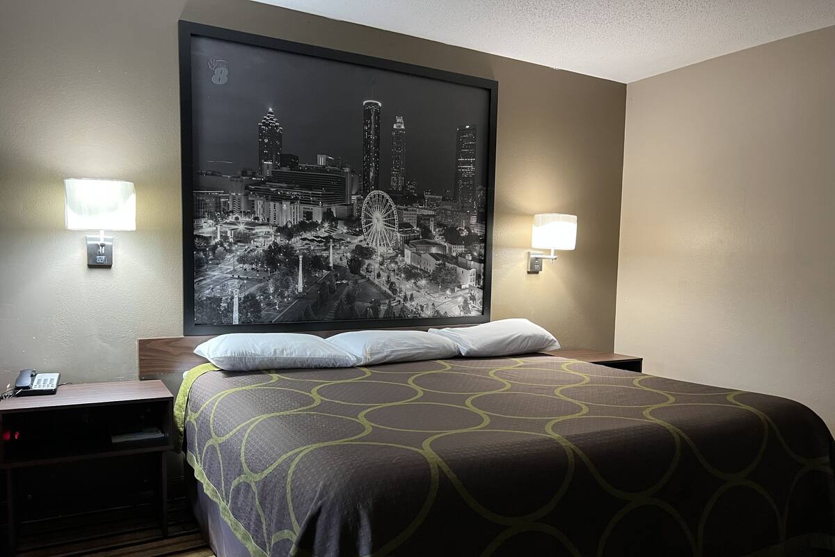A hotel room with a large bed, two bedside lamps, a nightstand with a phone, and a cityscape artwork on the wall.