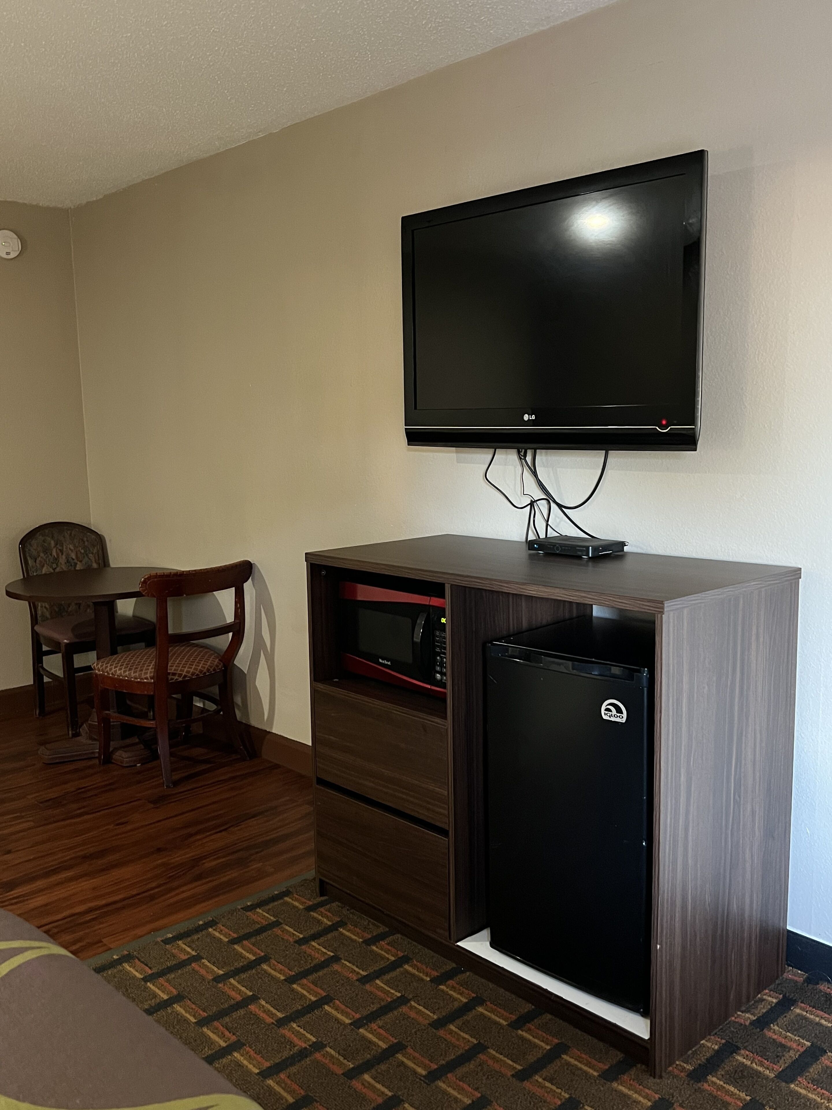 A hotel room with a flat-screen TV mounted on the wall, a wooden cabinet with a microwave and a mini-fridge, a wooden dining table with a chair, and a patterned carpet.