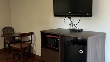 A hotel room with a flat-screen TV mounted on the wall, a wooden cabinet with a microwave and a mini-fridge, a wooden dining table with a chair, and a patterned carpet.