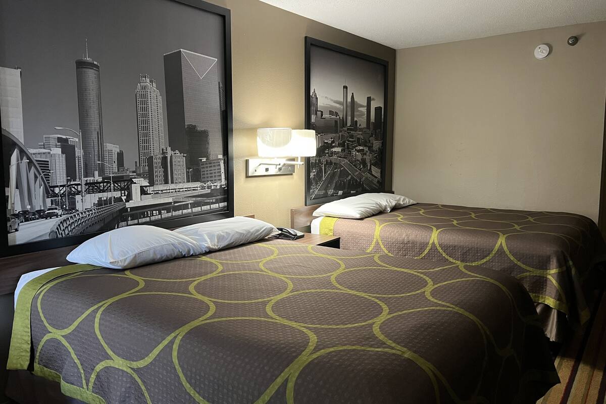 A hotel room with two beds, a large framed cityscape picture on the wall, a nightstand with a lamp, and a view of a city bridge through the window.