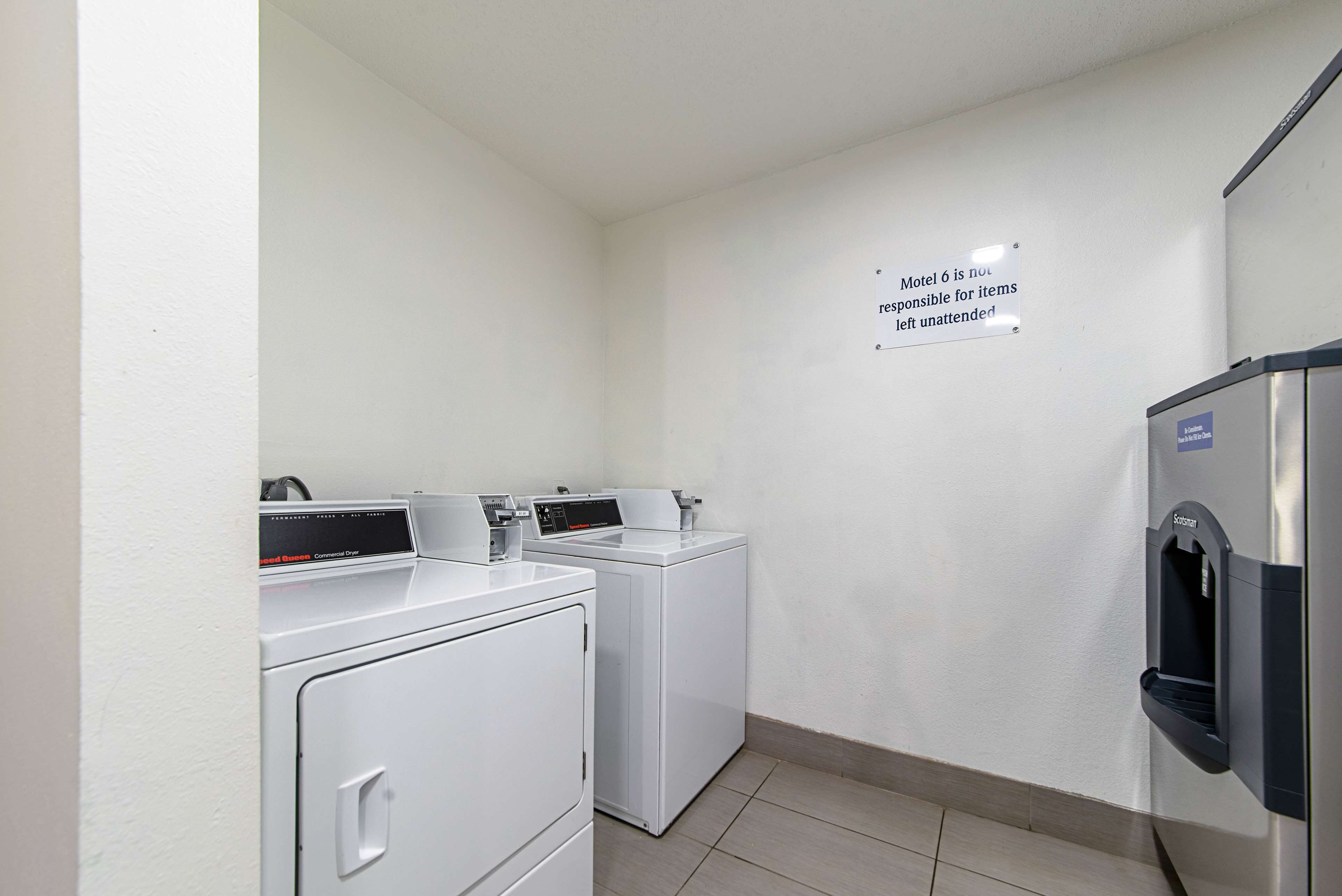 A laundry room with two front-loading washing machines, a wall-mounted dryer, and a water fountain. A sign on the wall states "Motel 6 is not responsible for items left unattended."