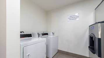 A laundry room with two front-loading washing machines, a wall-mounted dryer, and a water fountain. A sign on the wall states "Motel 6 is not responsible for items left unattended."