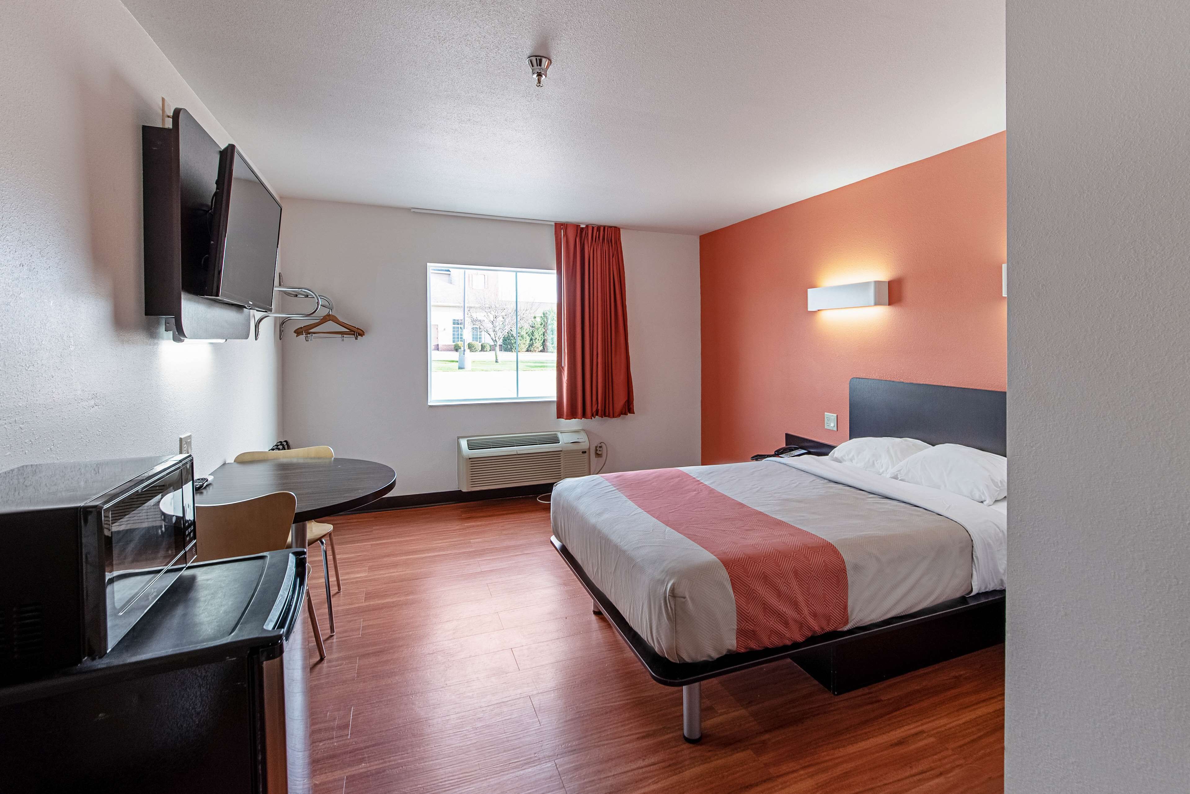 A hotel room with a bed, a desk, a chair, a TV mounted on the wall, and a window with curtains.