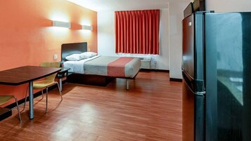 Quarto de hotel com cama, escrivaninha, cadeira, frigorífico e janela com cortinas.