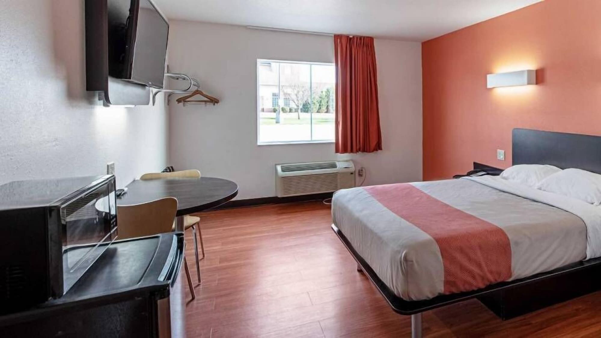 Ein Hotelzimmer mit einem Bett, einem Schreibtisch, einem Sessel, einem Fernseher, einem Fenster mit Vorhängen und einer Wandleuchte.