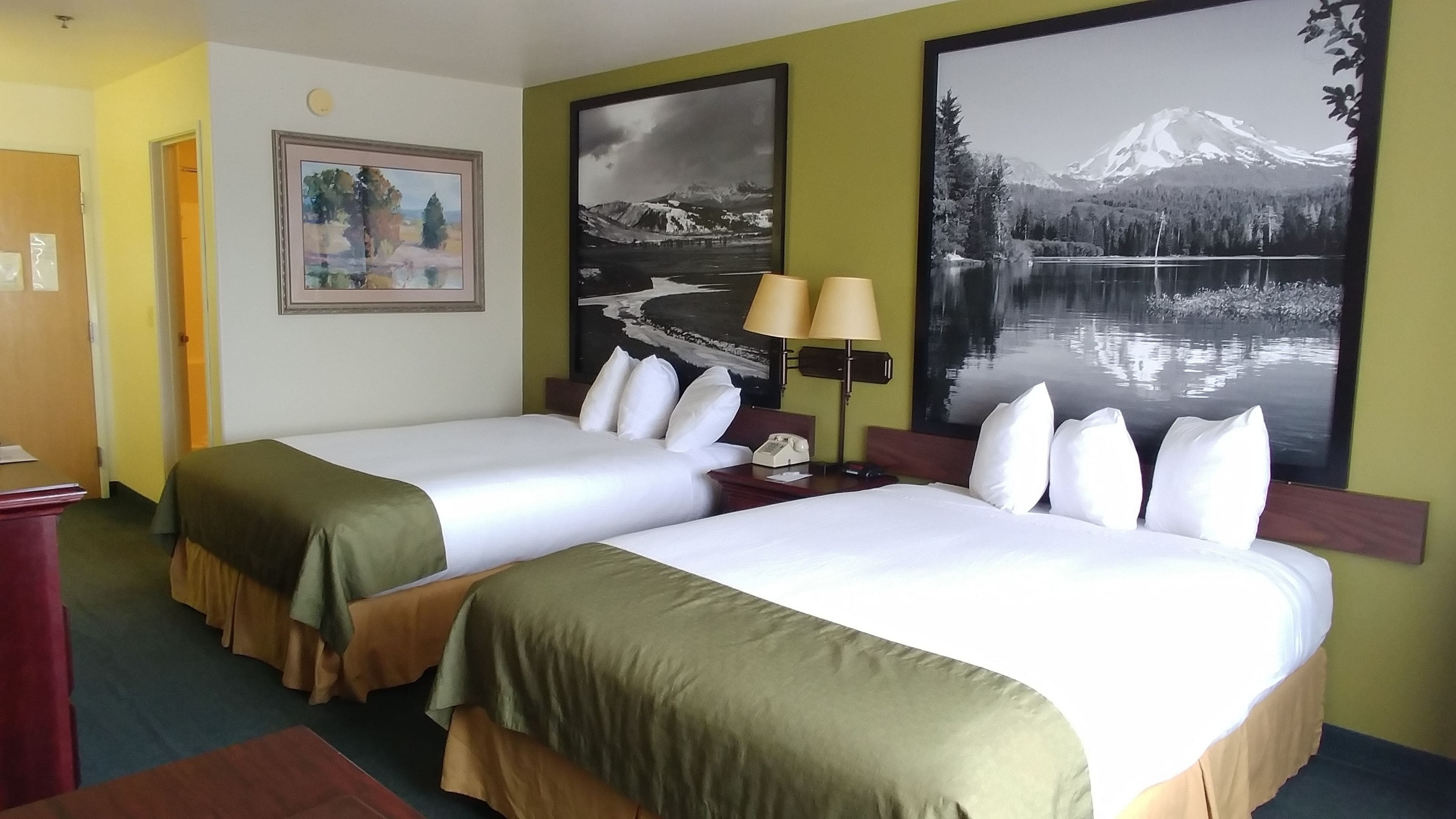 A hotel room with two beds, a desk, a lamp, and large landscape paintings on the walls.
