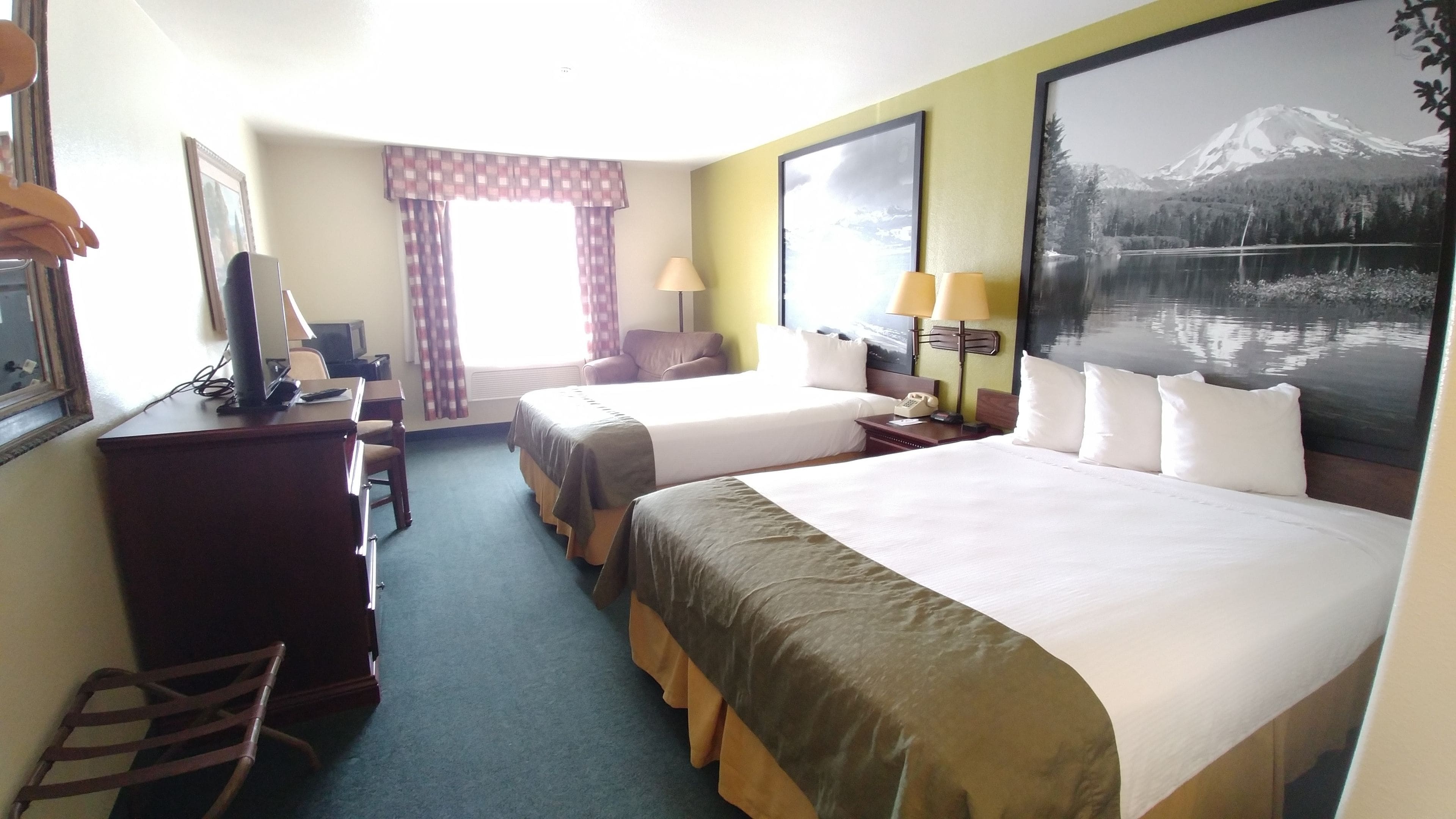 A hotel room with two beds, a desk with a chair, a TV, and a large window with a mountain view.