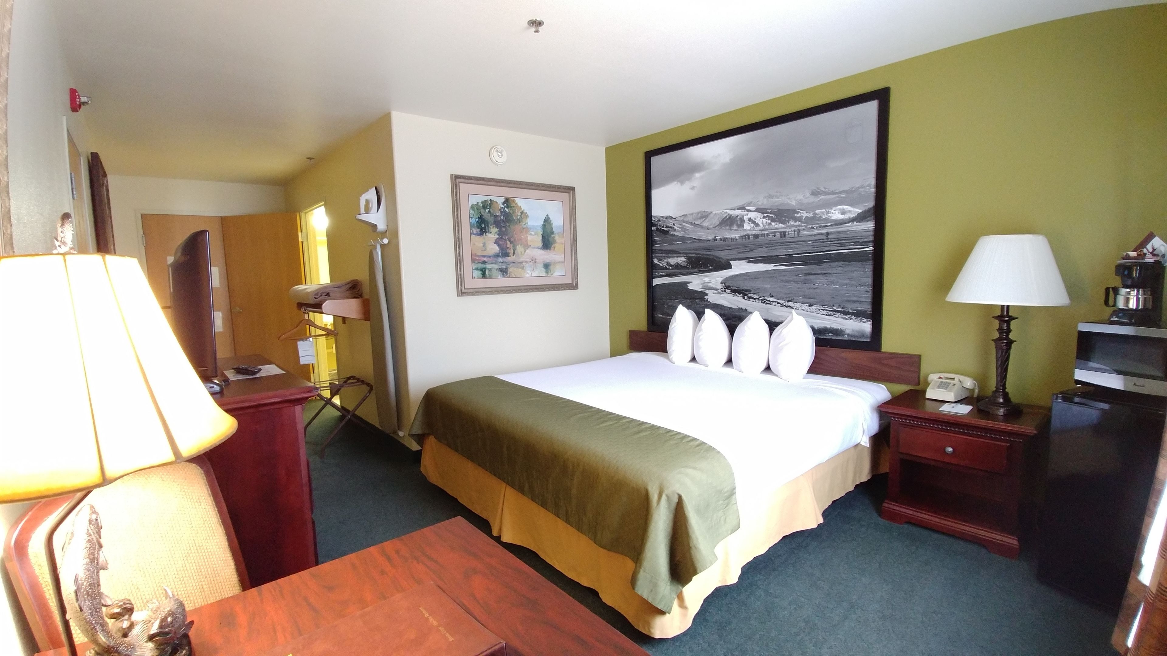 A hotel room with a bed, bedside tables, a desk, a lamp, a painting, and a view of a mountainous landscape.