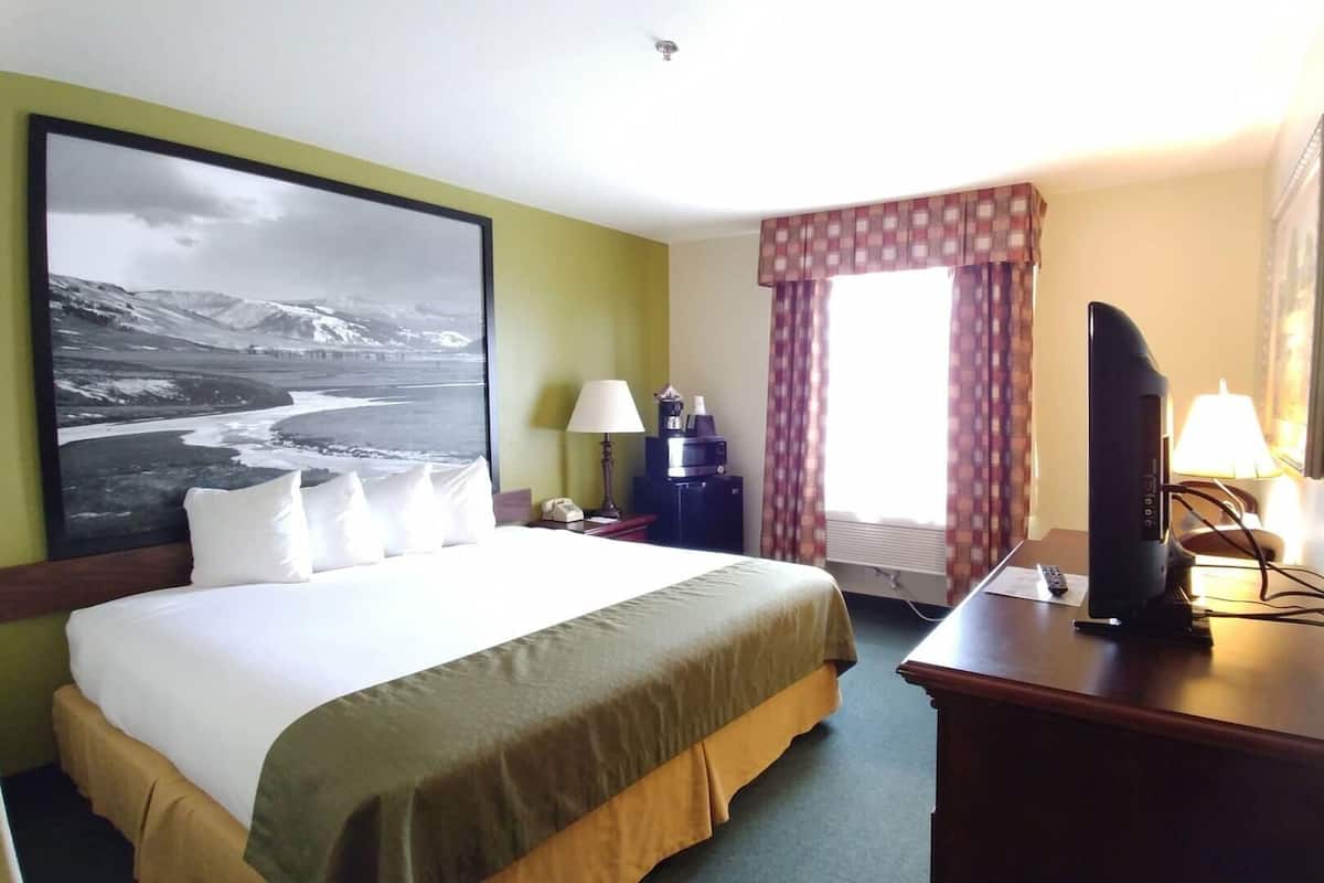 A hotel room with a bed, a desk with a computer, a TV, a window with curtains, and a painting on the wall.