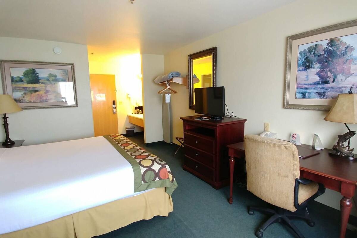 A hotel room with a bed, desk, chair, desk lamp, and a painting on the wall.