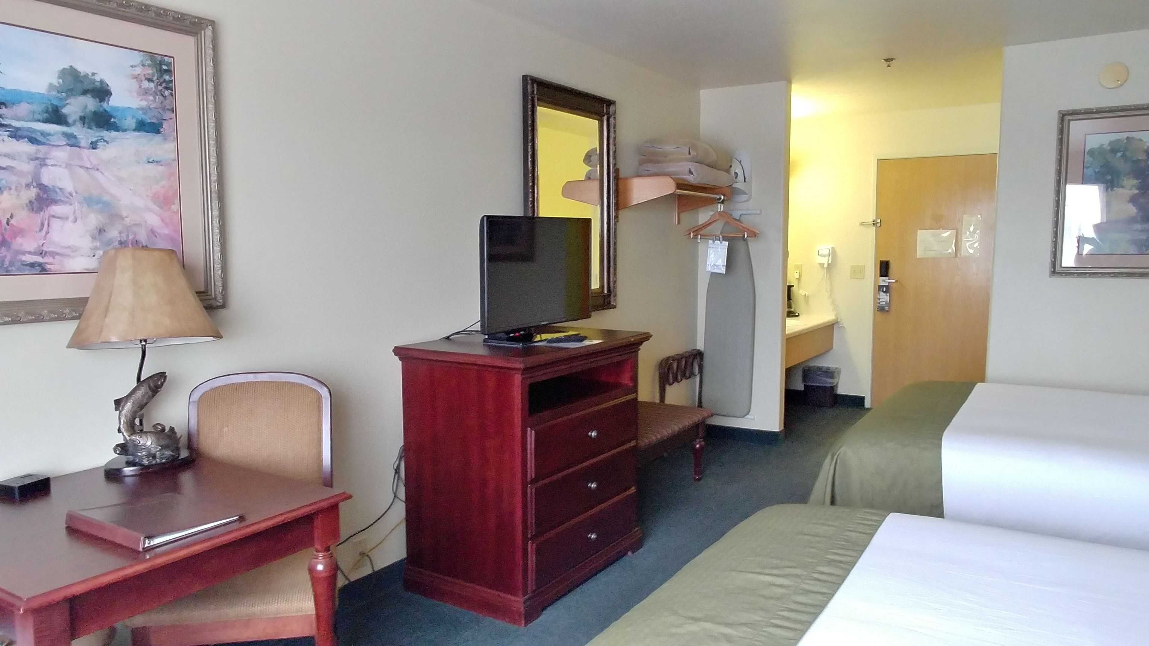 A hotel room with a bed, a desk with a lamp, a dresser, a television, and a mirror.