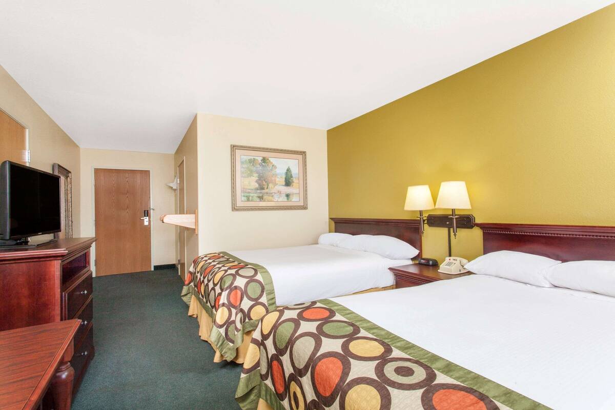A hotel room with two beds, a TV, a painting on the wall, and a desk with a lamp.