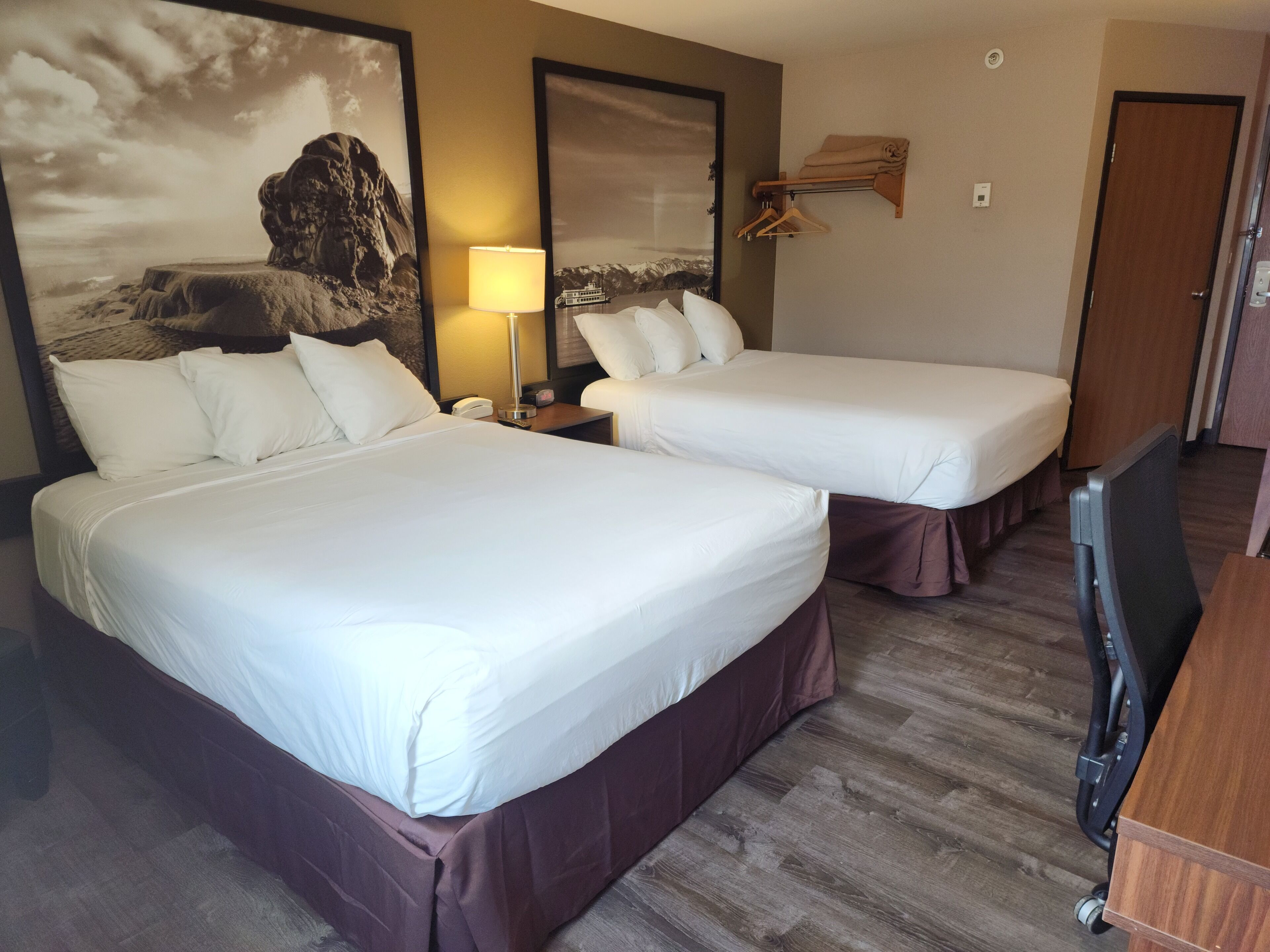 Habitación de hotel con dos camas, una mesita de noche, una lámpara, un estante de madera con toallas y un cuadro grande en la pared.