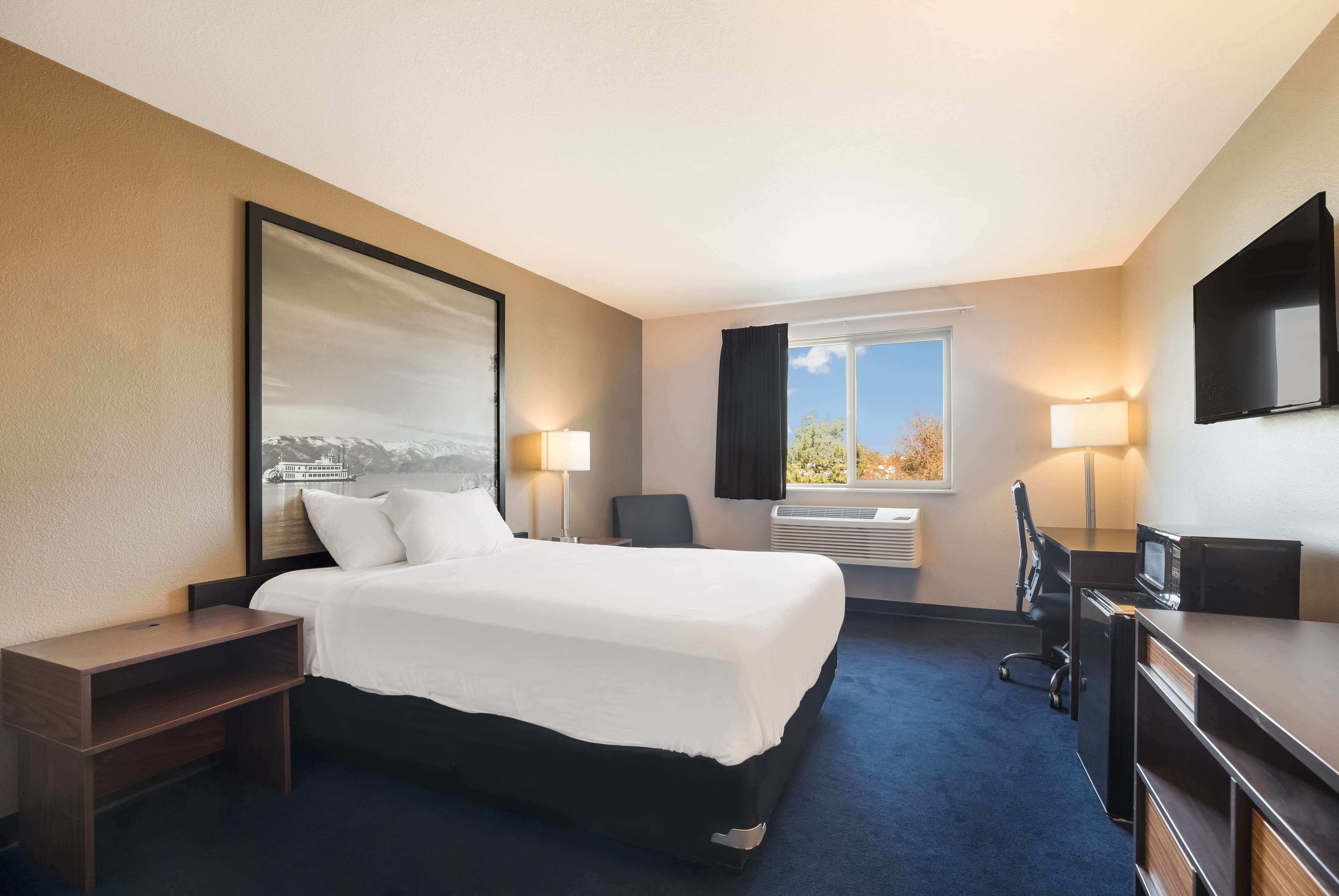 A hotel room with a bed, bedside tables, a desk, a chair, a TV, and a window with a view of trees.