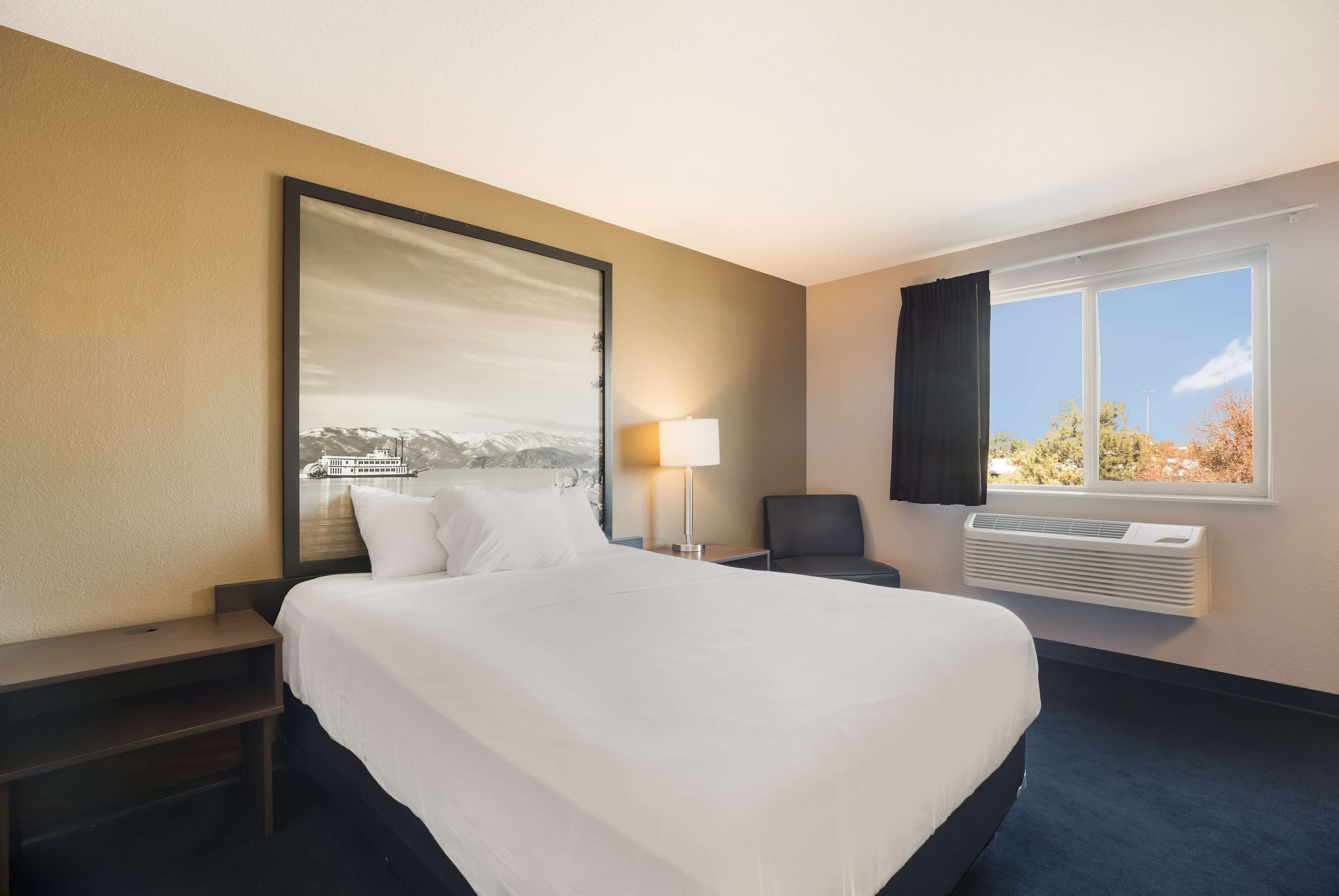 A hotel room with a large bed, a nightstand, a desk lamp, a chair, a window with a view of trees, and a wall-mounted air conditioner.