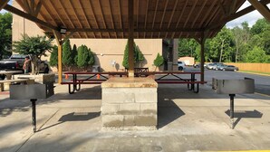 A covered picnic area with benches and tables, a stone fireplace, and a parking lot in the background. - Red Roof Inn PLUS+ Birmingham East - Irondale/ Airport (Birmingham)