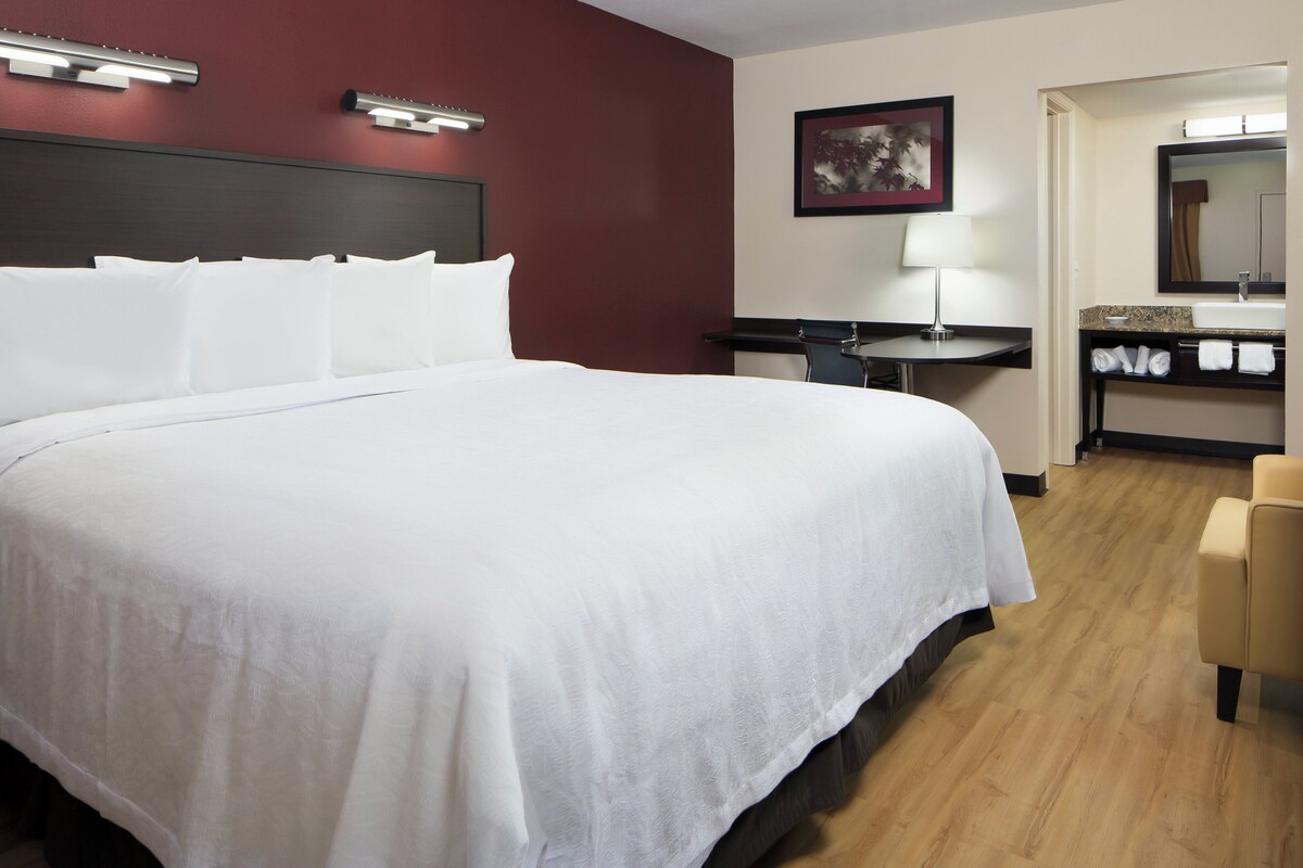 A hotel room with a large bed, a desk, a chair, a bathroom with a sink and mirror, and a painting on the wall.