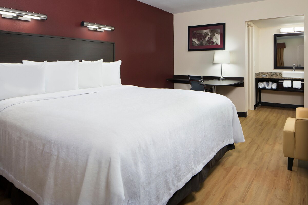Habitación de hotel con una cama grande, un escritorio, una silla, un baño con lavamanos y espejo, y un cuadro en la pared.