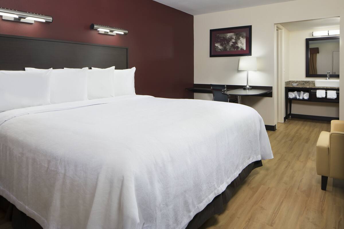 A hotel room with a large bed, a desk, a chair, a bathroom with a sink and mirror, and a painting on the wall.
