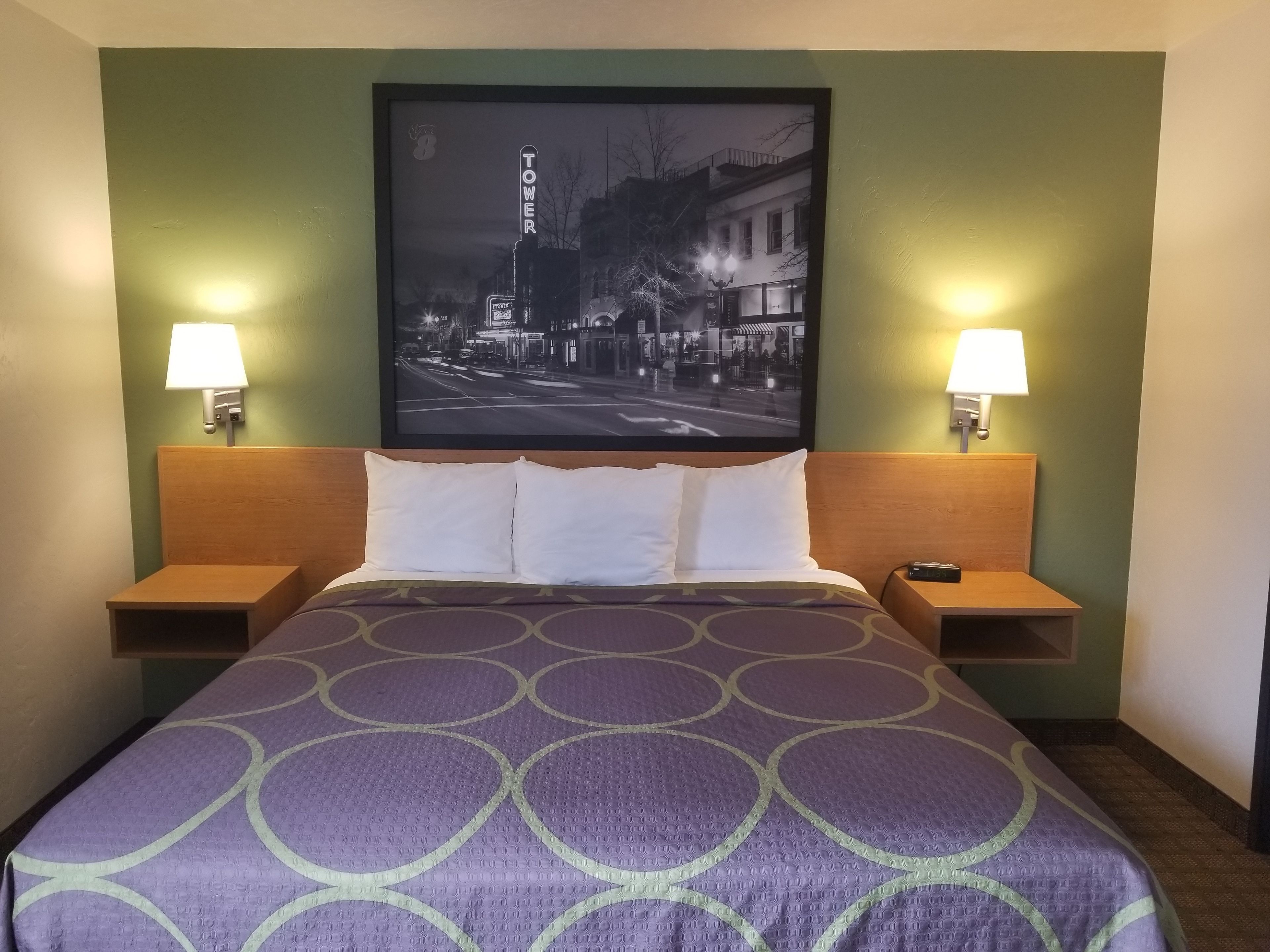 A hotel room with a bed, two bedside tables, two wall-mounted lamps, and a framed cityscape picture above the bed.