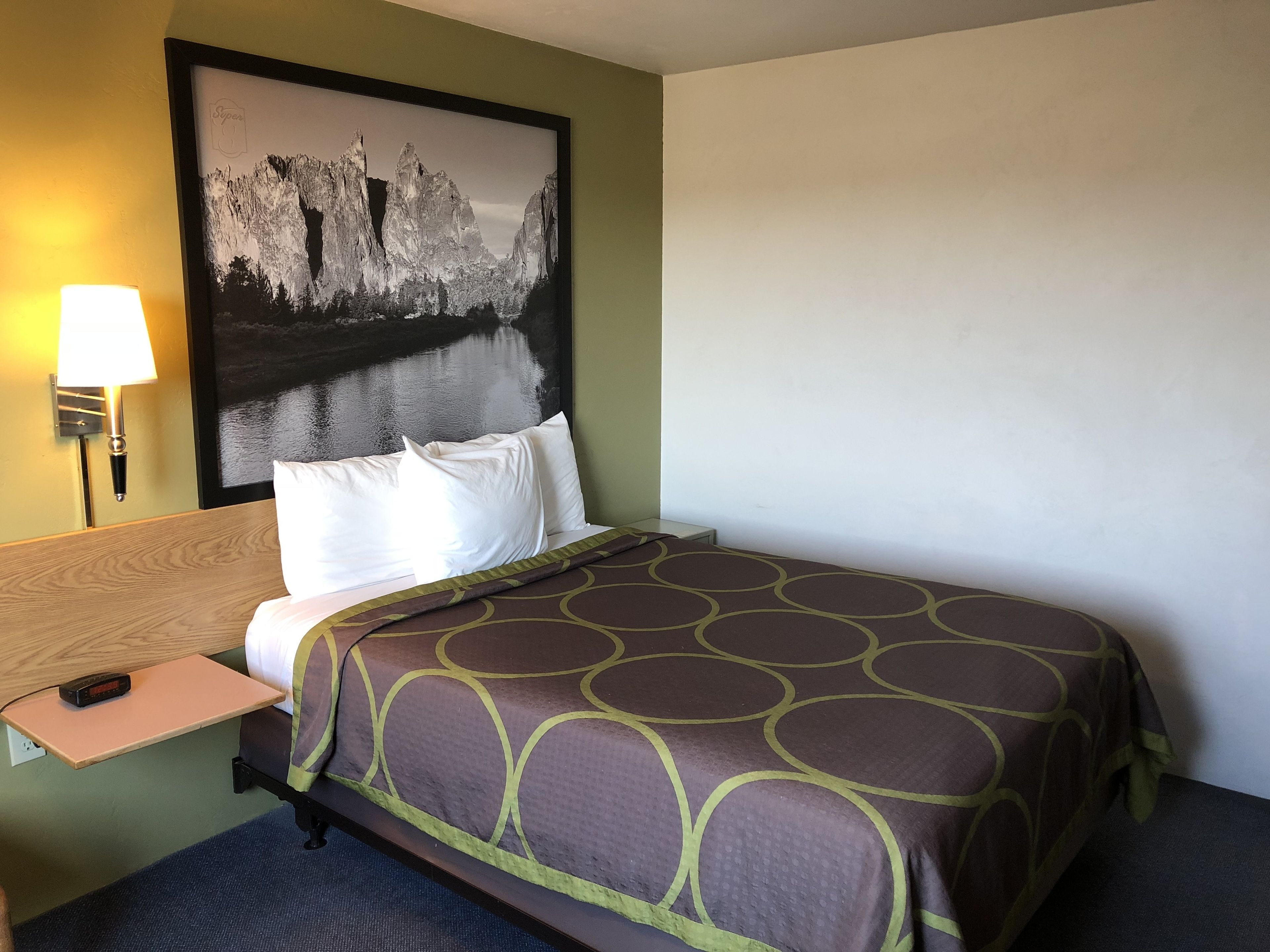 Habitación de hotel con cama, mesita de noche, lámpara y un cuadro grande en la pared.