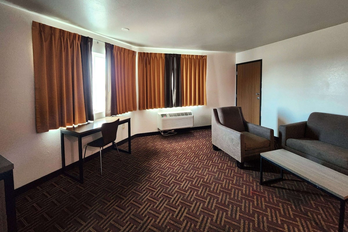 Ein Hotelzimmer mit einem Bett, einem Schreibtisch, einem Sessel und einer Couch. Es gibt ein Fenster mit Vorhängen, eine Klimaanlage und eine Tür.