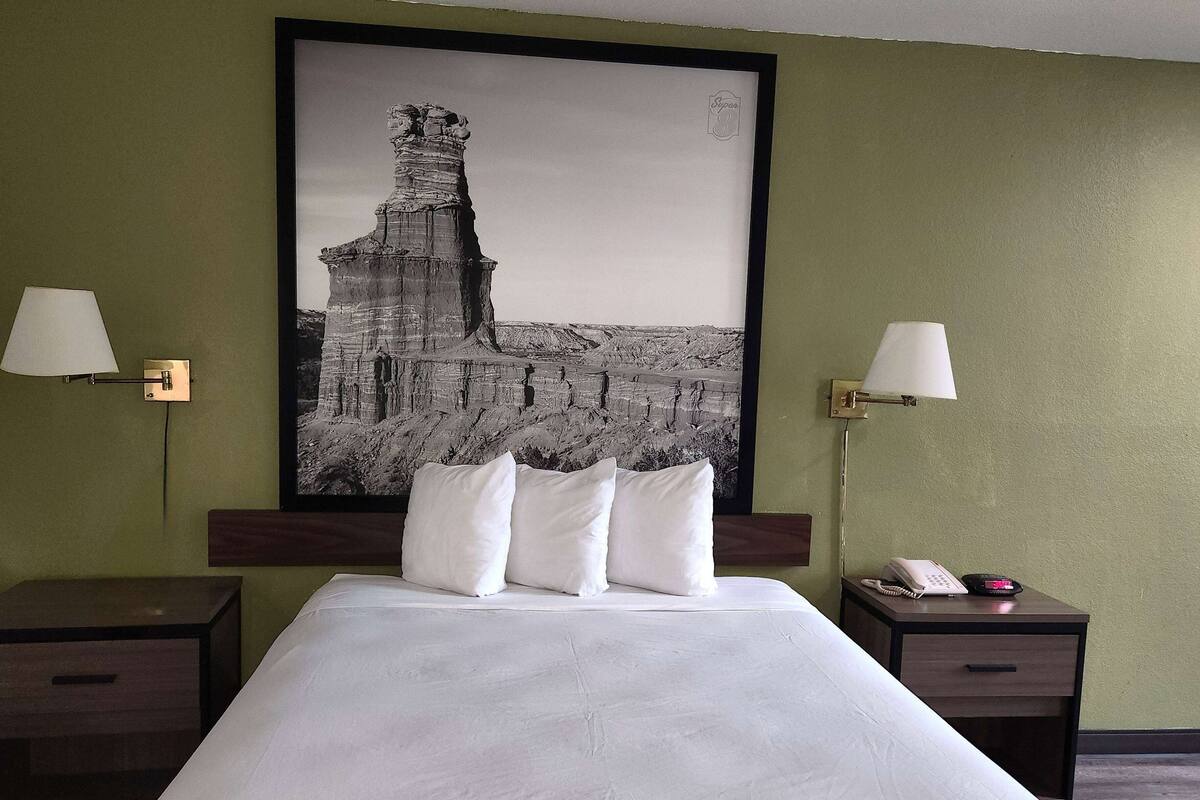 Ein Hotelzimmer mit einem großen Bett, zwei Nachttischen mit Lampen, einem gerahmten Schwarz-Weiß-Foto an der Wand und einem kleinen Tisch mit einem Telefon und einem roten Gegenstand.